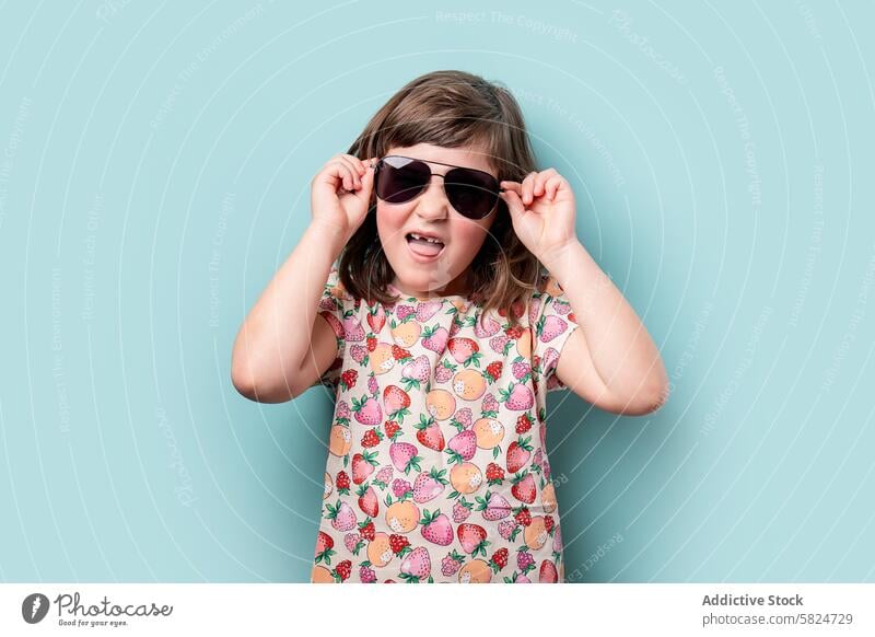 Young girl smiling and adjusting her sunglasses child smile fashion playful teal background young joyful fruit pattern summer style cute cheerful happy kid