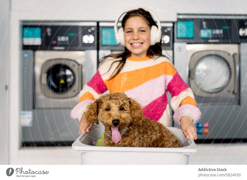 Preteen girl and dog having fun in laundry room preteen smiling basket headphones playful looking at camera indoor washing machines smile striped sweater joy