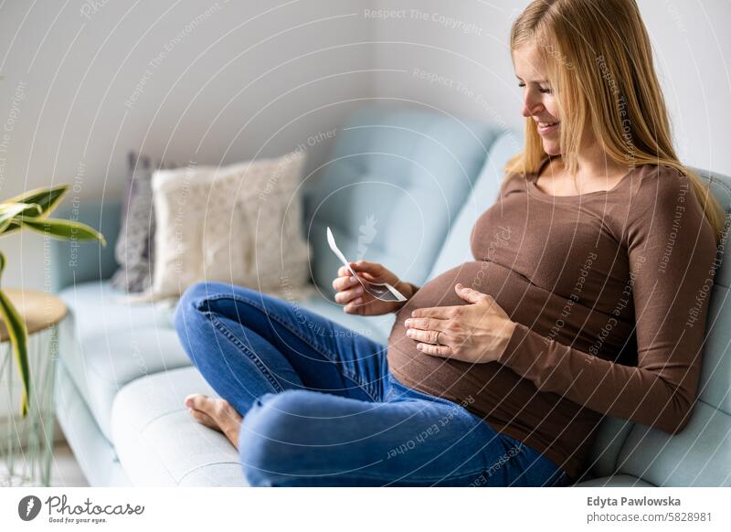 Pregnant woman sitting on sofa at home, looking at the ultrasound scan photo of her baby pregnancy pregnant adult anticipation awaiting belly birth body care