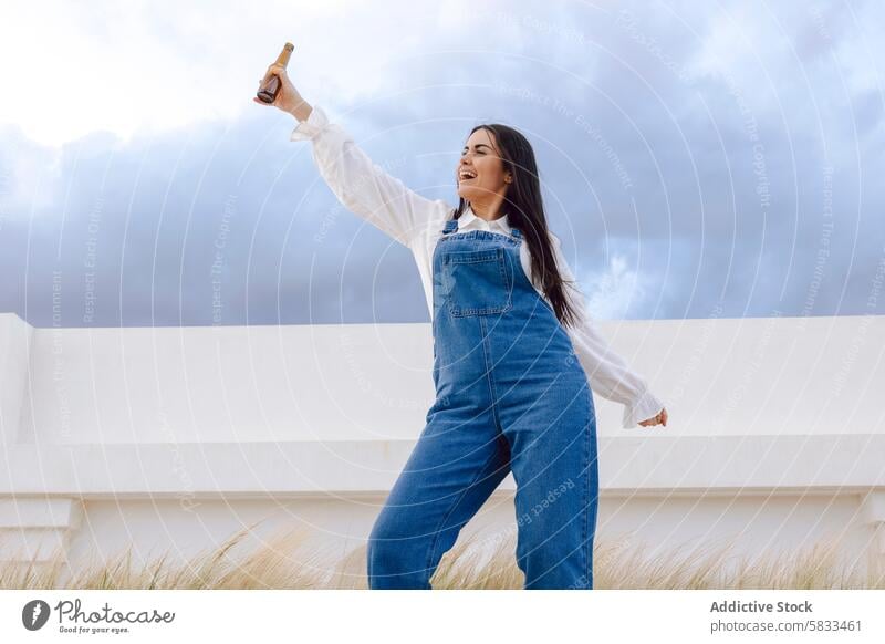 Joyful girl celebrating with beer outdoors woman bottle toast celebration joy freedom clouds sky daytime happiness cheerful expression arm raised overalls denim