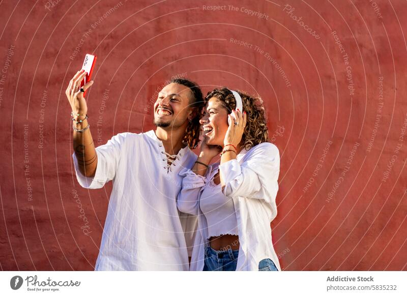 Young couple takes selfie with a smartphone against a red wall background young cheerful smiling joyous casual outfit white playful happy summer sunny day