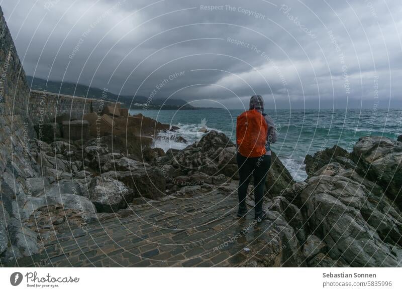 Tourist standing at the coast near the medieval harbor wall at the mediterranean sea on a cloudy rainy day, Cefalu, Sicily, Italy travel town tourist rock