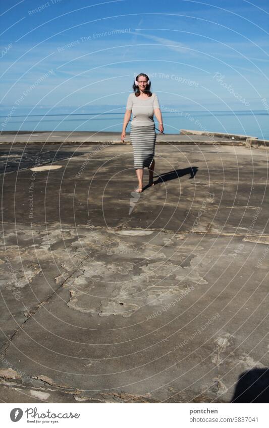 a woman with headphones walks across a concrete slab by the sea. happy, relaxing, unwinding. listening to music, listening to a podcast. Ocean holidays