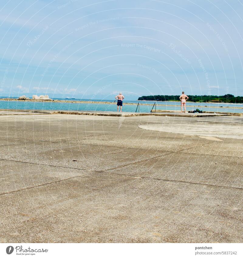 a man and a woman stand in the same posture with a view of the sea. a couple at a distance from each other. Posture Couple Woman Man Swimwear Ocean Stand