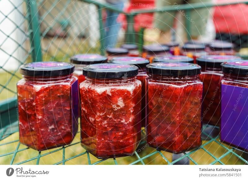 Collection of hexagonal jars with black lids filled with homemade strawberry jam or preserves in a grid-like pattern, wire mesh surface. The contents are bright red, with chunks of fruit and a slightly frothy appearance at the top, recently filled and p...