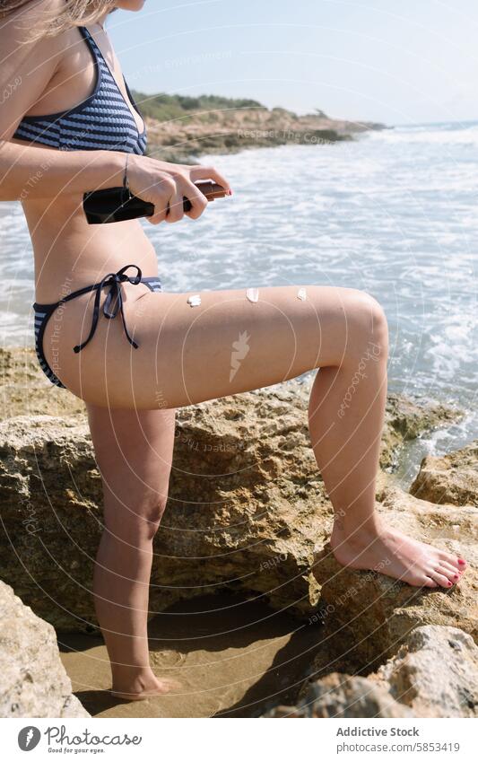 Woman Applying Sunscreen on Serene Beachside woman sunscreen application beach sea rocks bikini sunbathing tranquil sunny isolated coastline anonymous shore