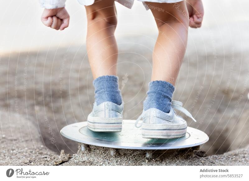 Child standing on a metal shovel on a sandy surface child feet blue sock white sneaker balance toddler close-up footwear texture gritty outdoor play adventure