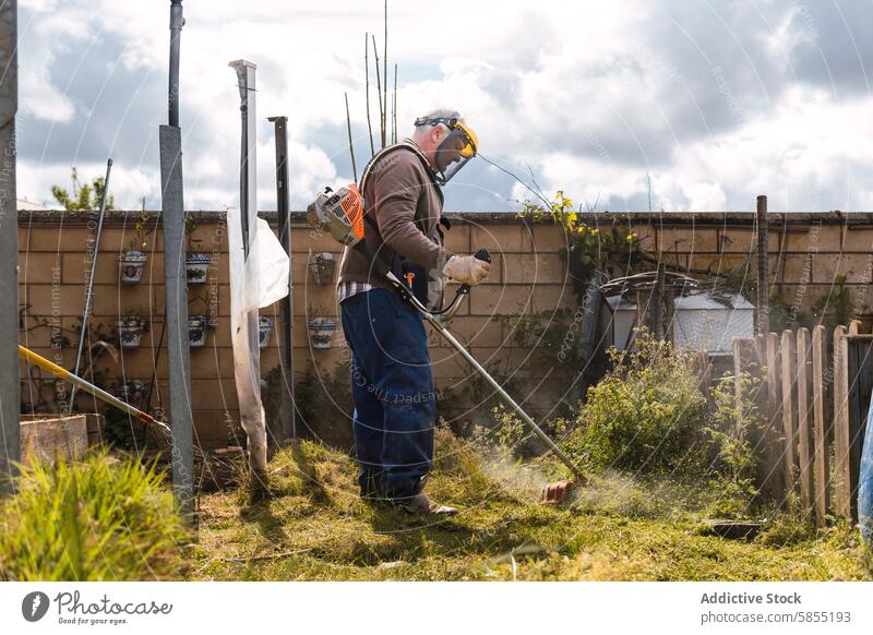 Man trimming overgrown grass with a brush cutter in backyard man garden work safety gear clouds sunny protective equipment outdoor task maintenance gardening