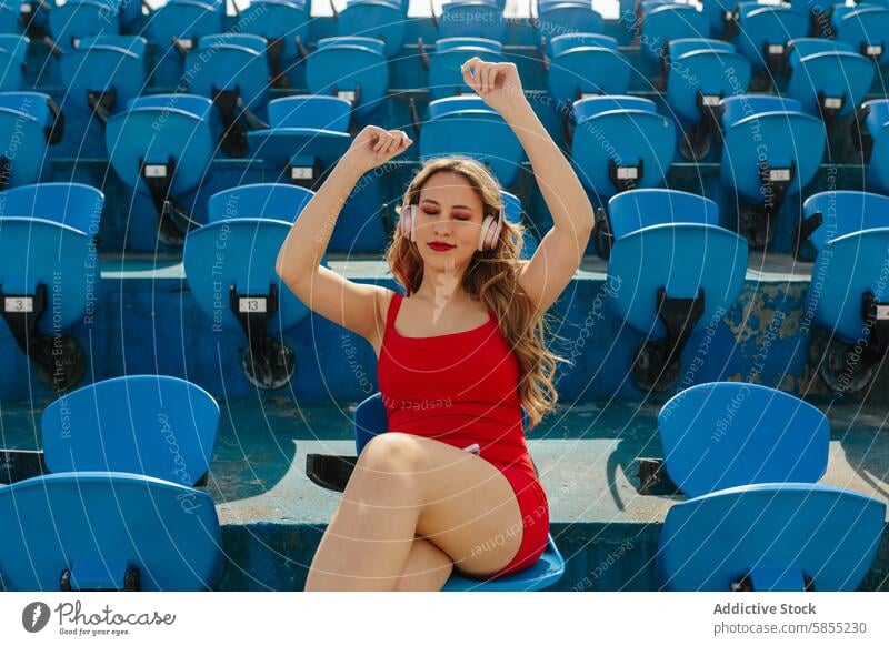 Young woman enjoying music alone in stadium seats headphones celebration red outfit blue seating entertainment leisure relaxation listening sitting outdoor