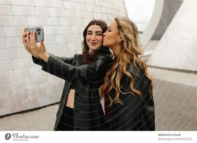 Two women taking a selfie with a modern background woman friend smartphone kiss cheek joy candid moment young happy sharing enjoyment urban fashion style