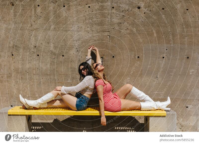 Stylish friends enjoying the sun on a park bench woman fashion trendy outfit relax pose bright yellow texture concrete sunlight summer leisure style outdoor fun