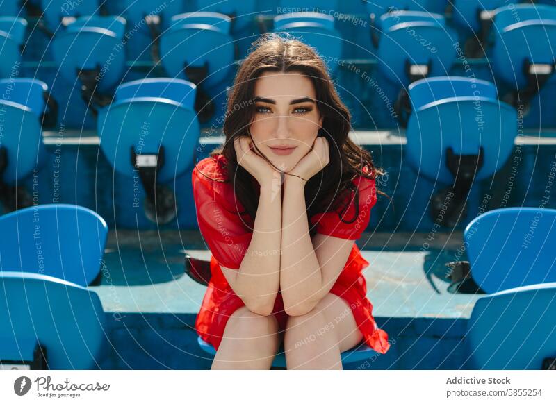 Young woman in red sitting alone in stadium seats dress gaze vibrant camera young fashion outdoor daytime cheerful thoughtful relaxed solitude individual blue