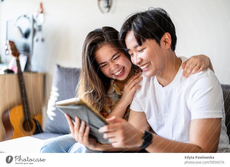 Happy young couple using digital tablet together on the sofa at home asian real people fun enjoying young adult authentic cheerful confident female happy