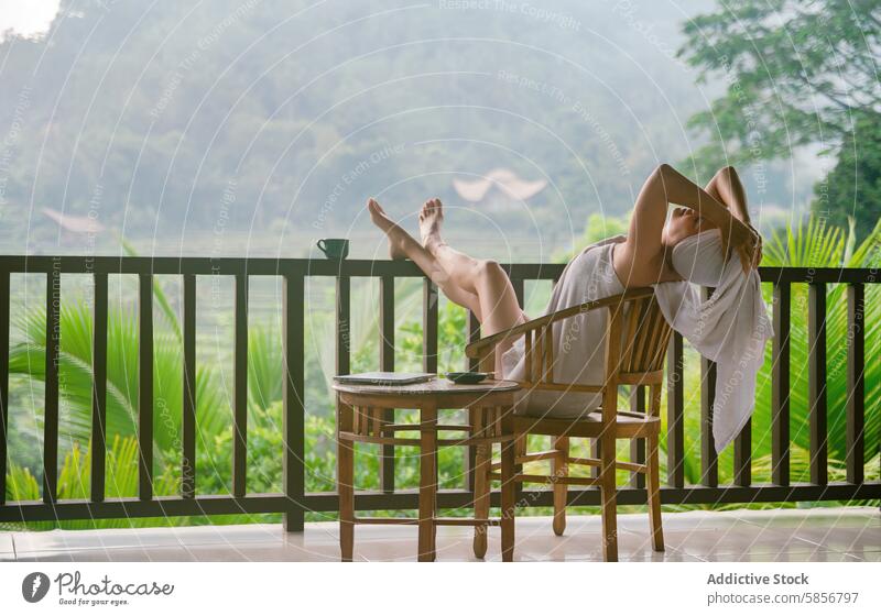 Woman Relaxing on Tropical Balcony Overlooking Misty Mountains woman relax balcony mountain mist view greenery serene tropical wooden chair feet up lush foliage