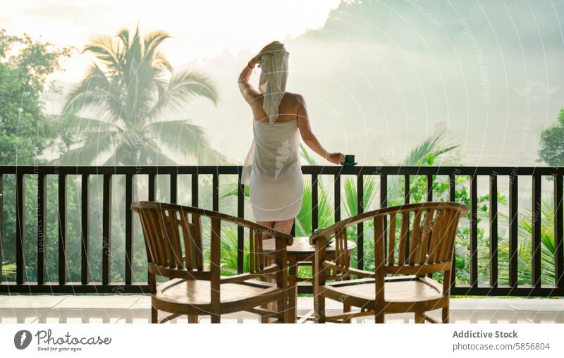 Woman relaxing on a tropical balcony with morning coffee woman mist view serenity towel hair nature outdoor furniture wooden chair rail palm tree cup green