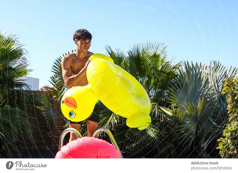 Young man with inflatable duck at poolside summer toy young shirtless holding yellow leisure outdoor looking away sunny palm trees swim shorts recreation fun