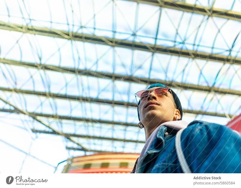 Young man in denim jacket under a geometric glass roof young stylish sunglasses sky blue urban fashion modern architecture structure outdoor day casual