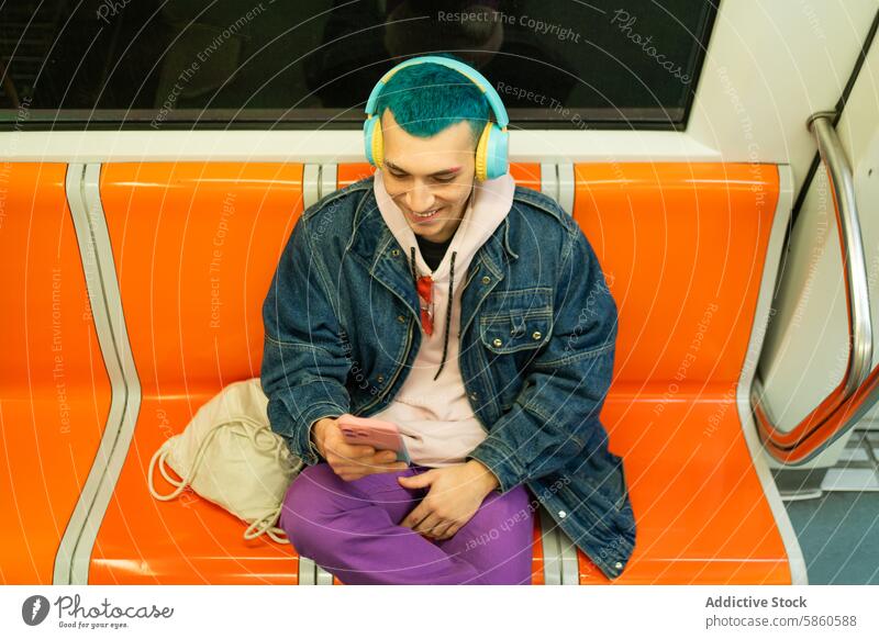 Young man with blue hair enjoying music on subway ride young headphone smartphone cheerful denim jacket purple trouser public transport seating absorbed joyful