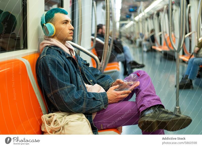 Young man with blue hair enjoying music on subway ride young headphone smartphone cheerful denim jacket purple trouser public transport seating absorbed joyful