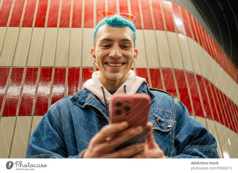 Young man with blue hair using smartphone in subway smile young red tile wall joyful denim jacket eyeglasses happy urban lifestyle casual technology