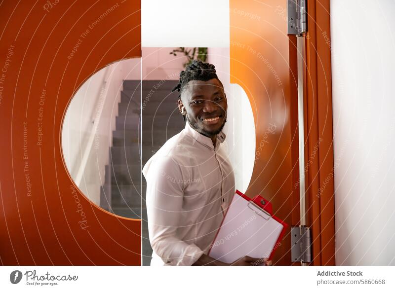 Confident young businessman with clipboard by office door inviting smiling professional cheerful open camera corporate welcoming confident workplace indoor