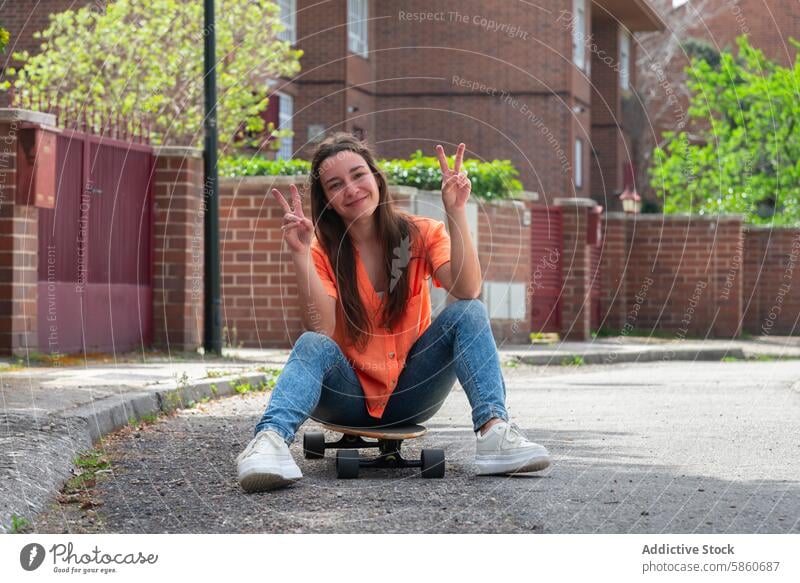 Smiling young woman sitting on skateboard making peace signs smile orange shirt jeans happiness suburban sidewalk outdoor leisure fun casual posing spring