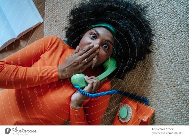 Comical 50s housewife talking on a vintage phone woman black telephone surprise reaction floor looking at camera orange sweater green headband indoor carpet