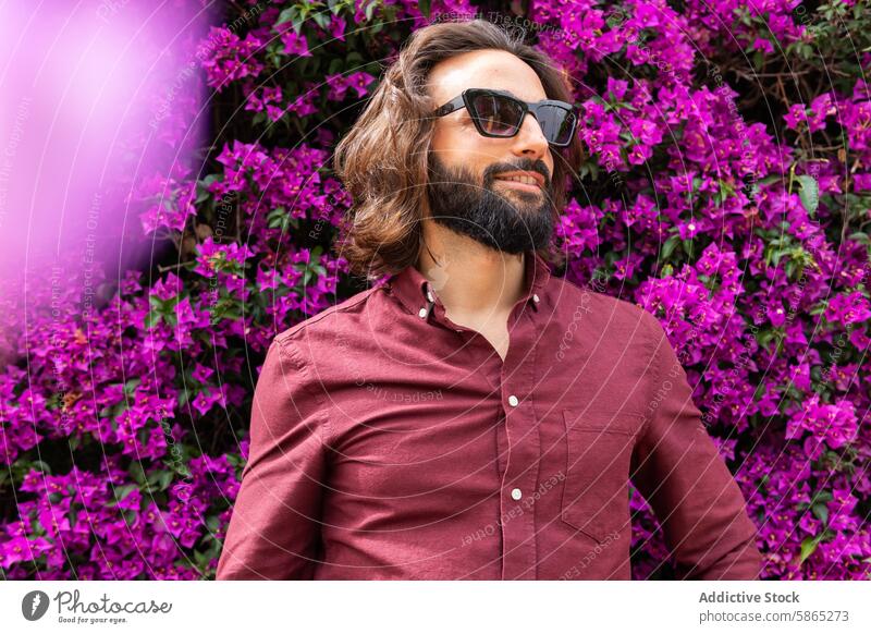 Stylish man with sunglasses posing against purple flowers beard casual style fashion trendy hip vibrant backdrop poblenou lifestyle outdoors daylight natural