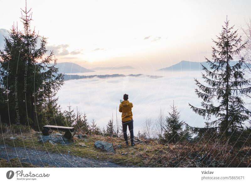 Adventurer takes photos on his mobile phone to capture the sunset and the clouds below. Capturing memories with technology. Beskydy mountains, Czech Republic