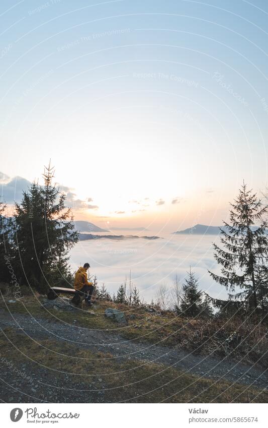 adventurer sitting on a wooden bench watches the clouds rolling in below him and the orange-yellow sunsets. The man contemplates and meditates. Beskydy mountains