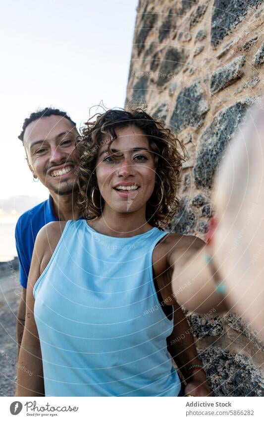 Close-up selfie of a joyful couple by a stone wall smile camera happiness woman dreadlocks direct gaze curly hair blue tank top outdoors caucasian cheerful