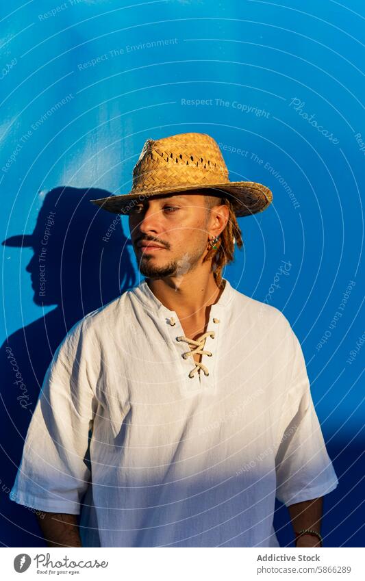 Man with dreadlocks and straw hat against blue wall man blue background profile shadow sunlight caucasian thoughtful looking away summer casual style vibrant