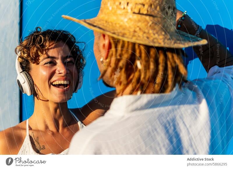 Smiling woman with headphones enjoying conversation outdoors smiling eye contact lively curly hair dreadlock straw hat blue background joyful talking casual