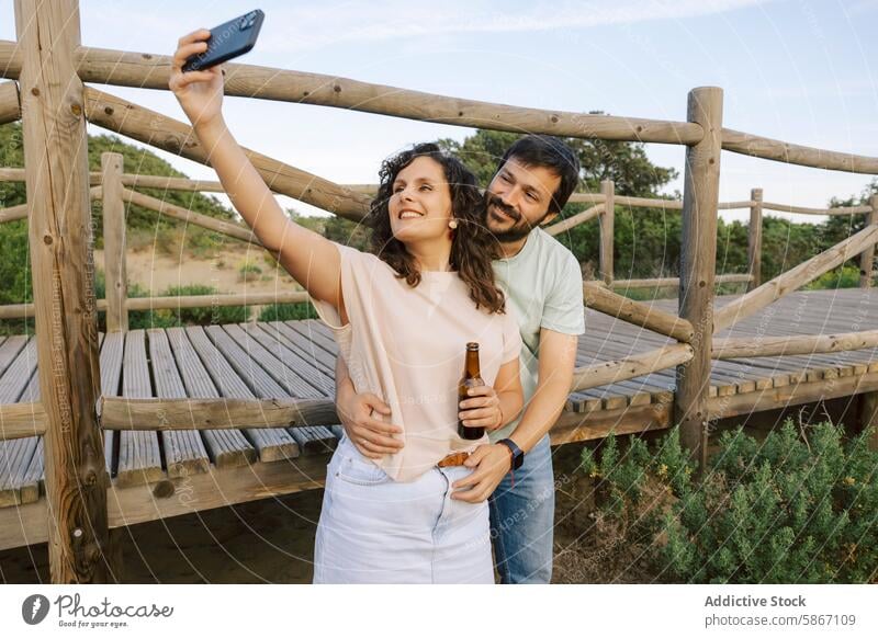 Couple enjoying beers and taking a selfie in nature couple smartphone outdoor wooden deck casual joyful peaceful man woman summer leisure relaxation drinking