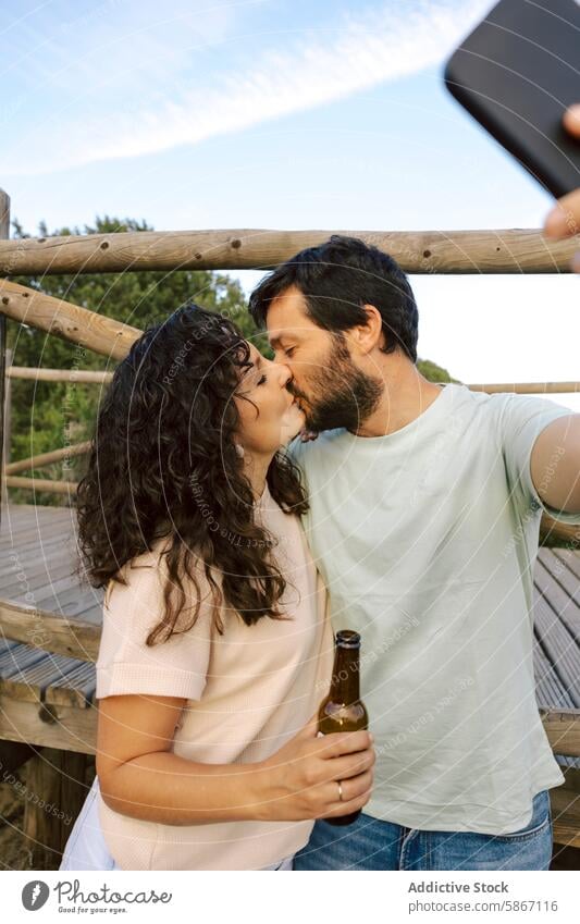 Couple kissing and taking a selfie with beer outdoors couple romantic casual moment intimacy wooded deck relationship love embrace mobile smartphone technology