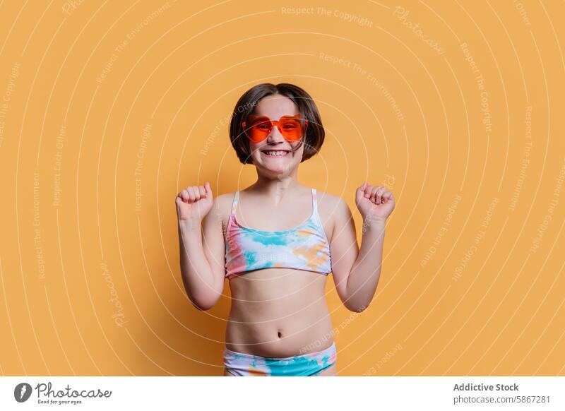 Joyful child in summer-themed attire on orange background joyful happy pose energetic spirit fun colorful tie-dye swimsuit heart-shaped sunglasses studio smile