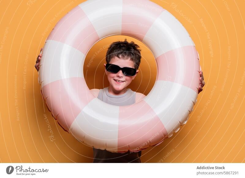 Boy with sunglasses smiling through a swim ring child summer studio theme boy smile orange background happiness joy playful fun leisure vacation youth striped