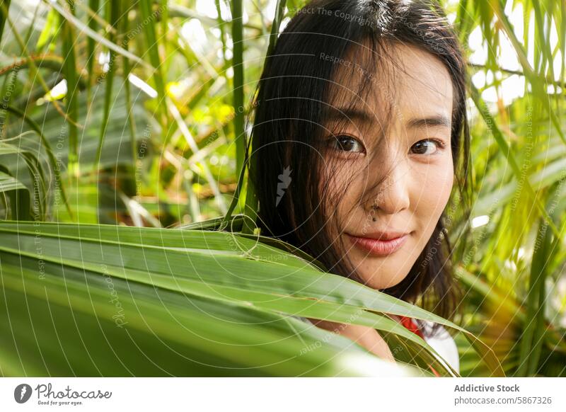 Young Asian woman peeking through lush green leaves asian close-up looking at camera vibrant playful serene expression nature outdoors young female eye contact