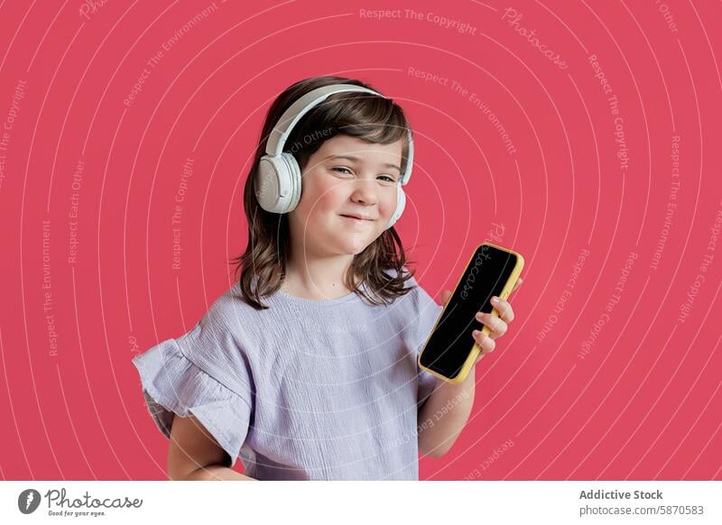 Young girl with headphones showing smartphone on red background studio child technology gadget mobile phone looking at camera music entertainment digital kid