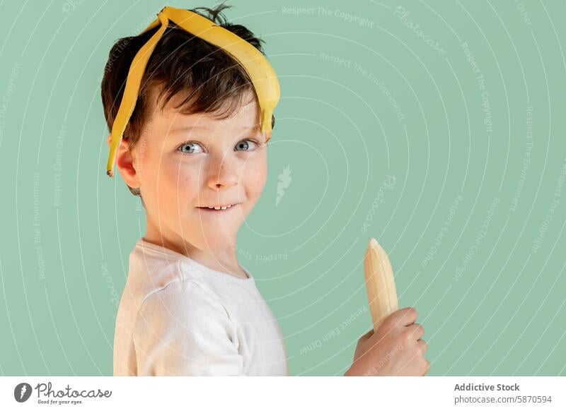 Young boy wearing a banana as hat and holding a peeled one studio child healthy snack fruit playful quirky costume creativity fun happy smile teal background