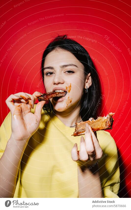 Young woman enjoys messy ribs against a red backdrop barbecue eating sauce finger face smudged yellow top enthusiastic background food casual meal happy