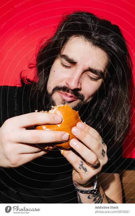 Man enjoying a delicious burger at a barbecue restaurant man eating enjoyment food meal tattoo hand red background bun meat juicy taste satisfaction appetite