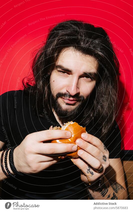 Man enjoying a juicy burger at a vibrant barbecue restaurant man young hair dark delicious bright red background relaxed inviting theme food eating savor tattoo