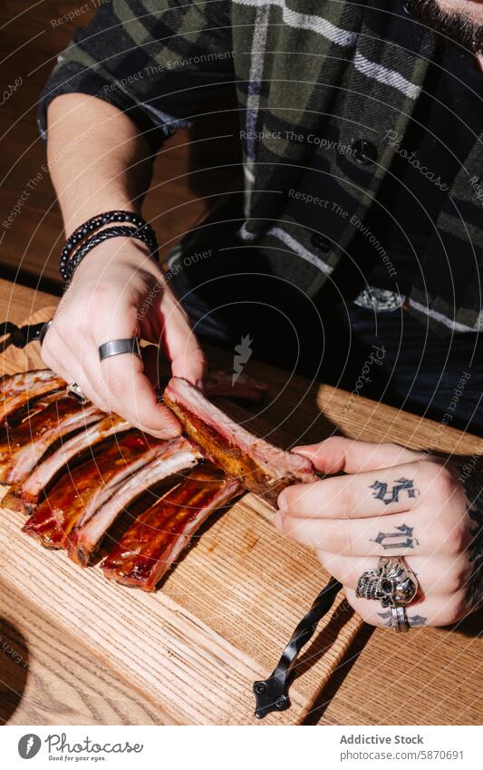 Man pulling ribs apart at a wooden table barbecue man meat plaid tattoo ring cooking food tender cutting board indoor meal dinner cuisine grilling eating hand