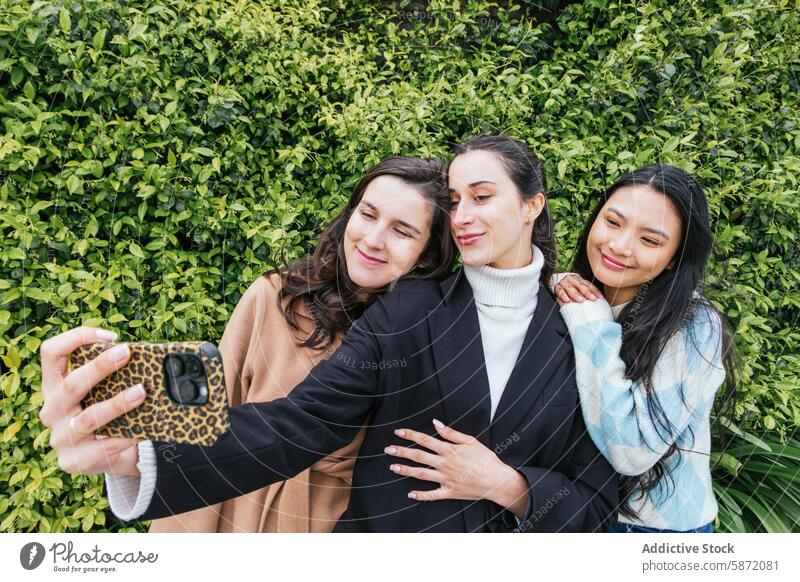 Three friends posing for a selfie with a green hedge background student female joyful friendship happiness together woman asian diversity smile camera phone