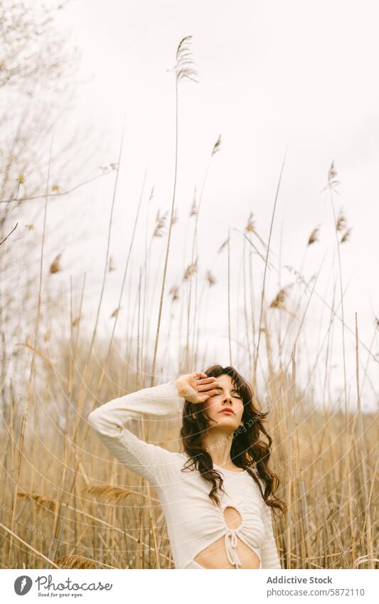 Young woman standing alone among tall grasses field tranquility introspection nature solitude young outdoor gaze pensive white top wilderness earthy tone