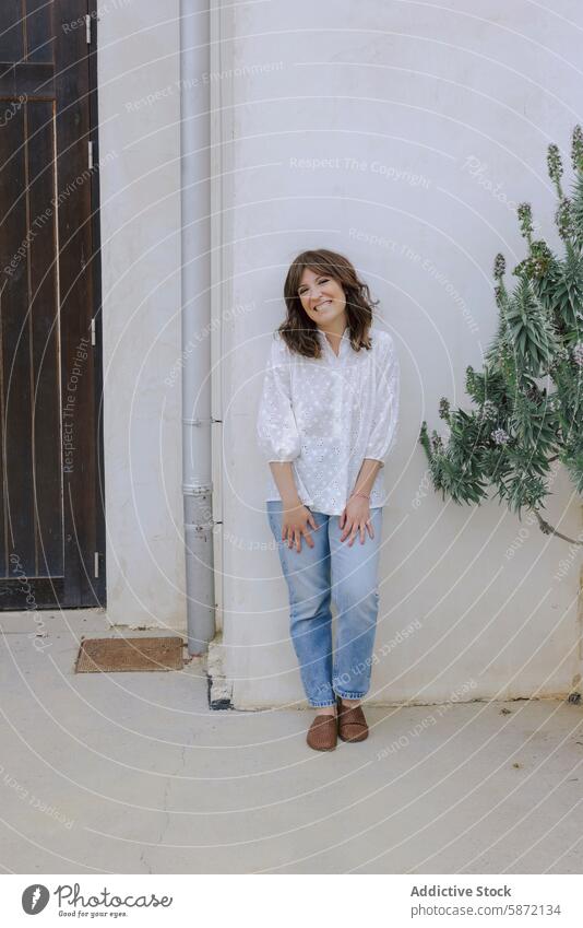 Cheerful woman in casual wear smiling against a textured wall smile joy happiness fashion style outdoor white blouse blue jeans standing leaning streetwear