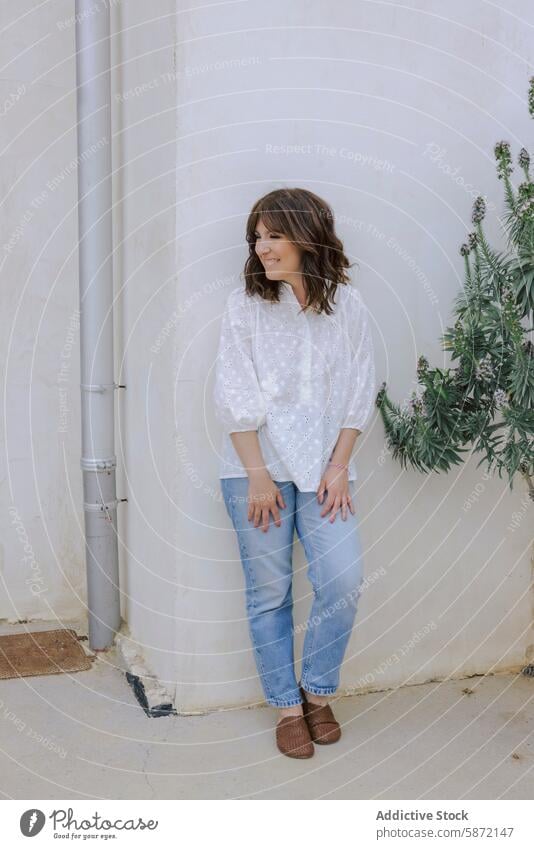 Smiling woman in casual outfit leaning against wall smile jeans blouse white blue happy joy serenity relaxed fashion style outdoor eyelet content cheerful