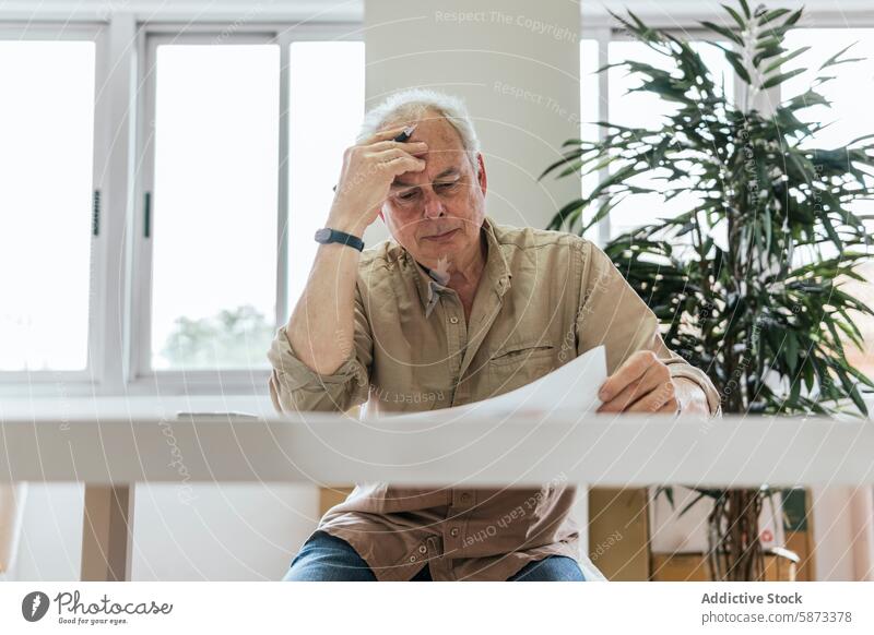 Pensive senior man looking at paperwork with concern in a bright room caucasian stress table white well-lit indoor plant document reviewing attention focused