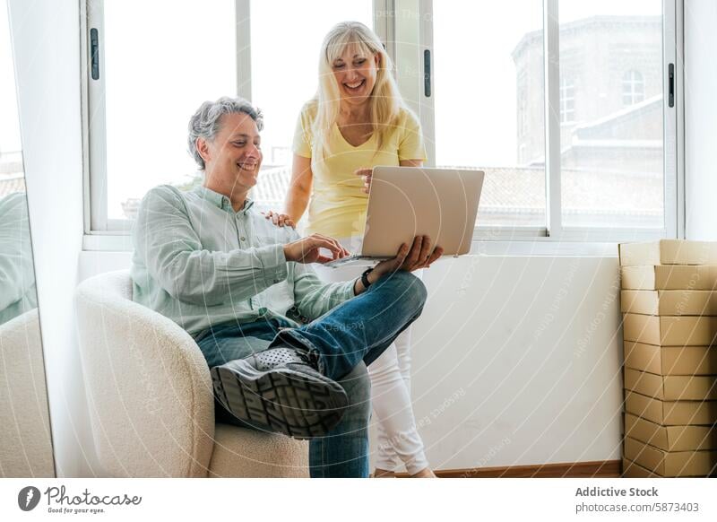 Smiling mature couple using laptop in bright room technology device gadget working cheerful sunlight casual indoors pointing looking at device working together